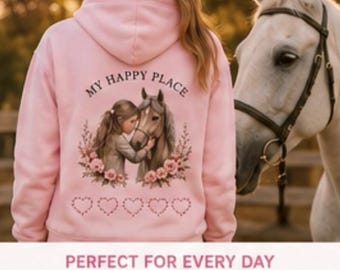 May include: A light pink hooded sweatshirt featuring a graphic on the back. The graphic shows a person embracing a horse, surrounded by flowers and the text "MY HAPPY PLACE" above, with a row of five heart outlines printed below the image.