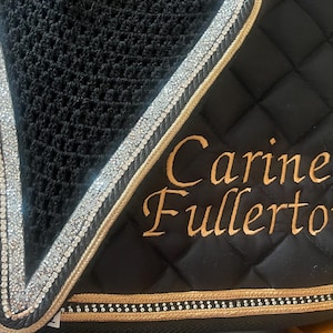 May include: Black quilted saddle pad with gold trim and silver rhinestone accents. The pad features the embroidered name "Carine Fullerton" in gold script. A black mesh material is visible at the top.
