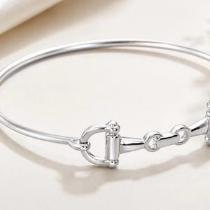May include: A polished silver-tone bangle bracelet featuring a central horse bit motif. The circular band is smooth and slender, connecting to a detailed metal bit design with two side rings and a horizontal bar, creating a cohesive metallic accessory.
