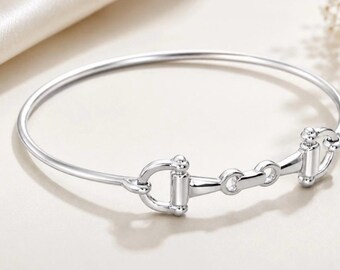 May include: A polished silver-tone bangle bracelet featuring a central horse bit motif. The circular band is smooth and slender, connecting to a detailed metal bit design with two side rings and a horizontal bar, creating a cohesive metallic accessory.