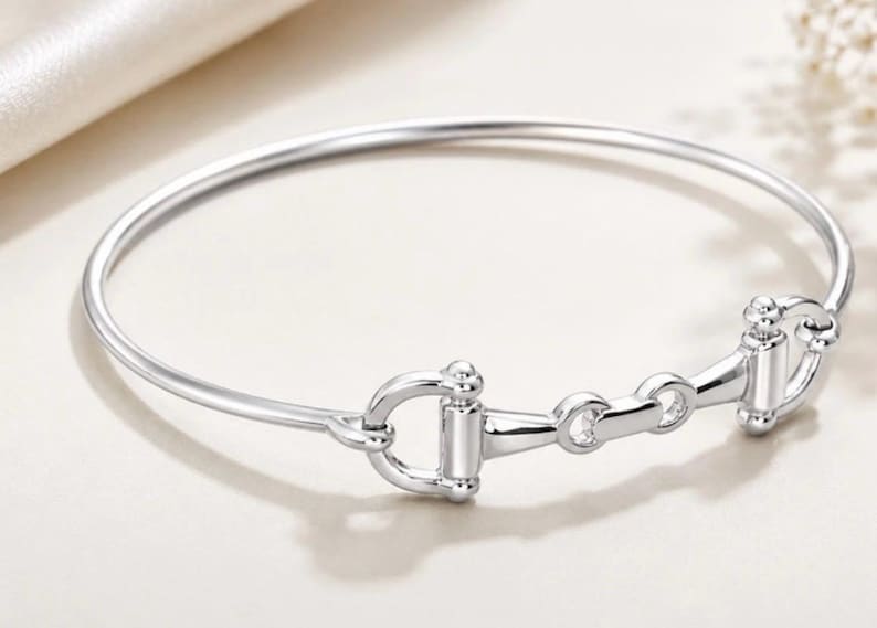 May include: A polished silver-tone bangle bracelet featuring a central horse bit motif. The circular band is smooth and slender, connecting to a detailed metal bit design with two side rings and a horizontal bar, creating a cohesive metallic accessory.