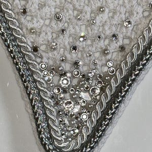 May include: A close-up view of a white crocheted item, adorned with sparkling rhinestones and small pearls. The edges are finished with silver and white braided trim, giving a luxurious and elegant appearance. The item has a delicate, handcrafted look.