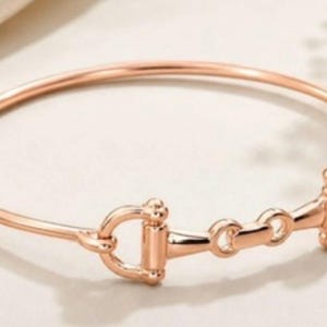 May include: A rose gold-toned bangle bracelet featuring a central horse bit motif. The bracelet has a smooth, polished finish and a circular band design that connects to the detailed metal hardware in the centre of the piece.