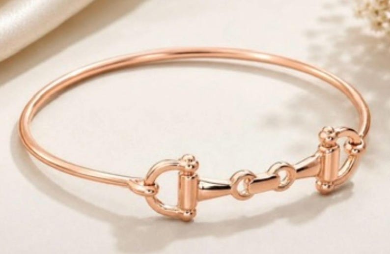 May include: A rose gold-toned bangle bracelet featuring a central horse bit motif. The bracelet has a smooth, polished finish and a circular band design that connects to the detailed metal hardware in the centre of the piece.
