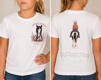 May include: A white short-sleeve t-shirt featuring a graphic on the front chest of a horse head with the text COWGIRL AT HEART and OLVOSSA. The back displays a graphic of a person riding a horse with the text ISLA and a row of heart outlines.