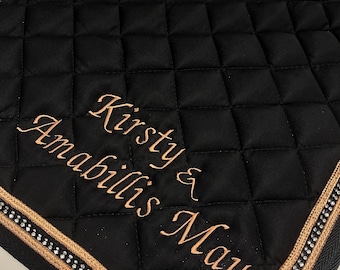 May include: Black quilted saddle pad with gold trim and silver accents. The pad features the embroidered names "Kirsty & Amabillis May" in a cursive font. The pad is designed for equestrian use.