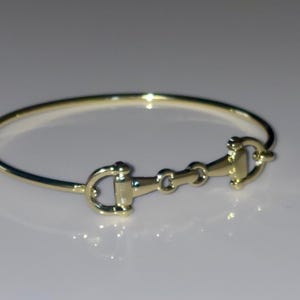 May include: A polished gold-tone bangle bracelet featuring a central horse bit motif. The circular band is connected to a horizontal metal piece that mimics the design of an equestrian bit, including two side rings and a central link.
