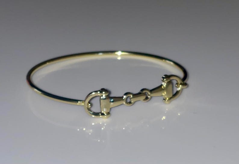 May include: A polished gold-tone bangle bracelet featuring a central horse bit motif. The circular band is connected to a horizontal metal piece that mimics the design of an equestrian bit, including two side rings and a central link.