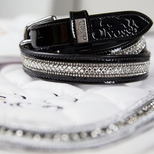 May include: A black leather belt with a glossy finish, embellished with sparkling rhinestones. The belt has a silver buckle and the brand name "Olivossa" embossed on the strap. It's displayed on a white quilted surface.