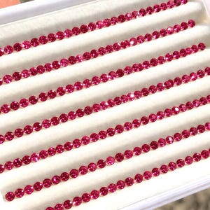 May include: A white display tray containing multiple rows of small, round, faceted pinkish-red gemstones. Each stone is uniform in size and shape, arranged in neat horizontal lines against a soft, white, padded velvet-like surface.