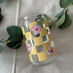 Op de afbeelding: Een transparante glazen beker met een geel en wit geruit patroon en roze bloemen met rode centra.