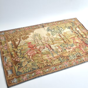 May include: A tapestry with a beige background depicting a hunting scene in a forest. The tapestry features a man on horseback, other figures, and a variety of trees and foliage. The tapestry has a brown, green, and blue border.
