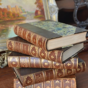 May include: A stack of five antique leather-bound books with gold accents and intricate designs. The books are arranged in a pyramid shape on a wooden surface.
