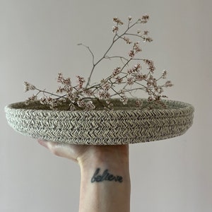 Può includere: Un vassoio rotondo intrecciato di colore beige con un motivo bianco e nero. Il vassoio è tenuto da una mano con un tatuaggio che dice "believe". Ci sono delicati fiori rosa nel vassoio.