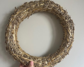 Può includere: Una ghirlanda circolare fatta di paglia secca intrecciata. La ghirlanda è di colore beige naturale e sembra fatta a mano. La ghirlanda è tenuta da una mano contro un muro bianco.