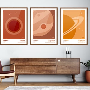 K&ouml;nnte beinhalten: Drei minimalistische Poster mit Illustrationen von Mars, Jupiter und Saturn. Jedes Poster zeigt ein anderes Farbschema und eine Nummer, die die Position des Planeten im Sonnensystem darstellt. Die Poster sind in braunem Holz gerahmt und an einer wei&szlig;en Wand aufgeh&auml;ngt.