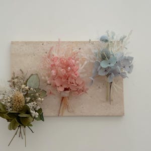 Può includere: Tre piccoli bouquet di fiori secchi nei toni del verde, rosa e blu sono disposti su una superficie beige rettangolare e strutturata. I bouquet sono legati con spago. Lo sfondo è un muro bianco.
