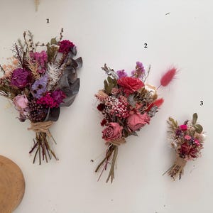 May include: Three bouquets of dried flowers in various shades of pink, purple, and white. The bouquets are arranged in a row from largest to smallest, with the largest bouquet on the left and the smallest bouquet on the right. The bouquets are tied with twine.