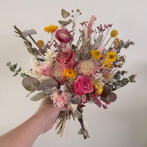 May include: A vibrant bouquet of dried flowers featuring a mix of colors including pink, yellow, and green. The arrangement includes roses, daisies, and eucalyptus leaves, held together with a natural twine.