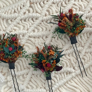 Puede incluir: Tres horquillas con arreglos de flores secas. Las flores son en tonos de naranja, rojo y verde. Las horquillas son negras y tienen una doble punta.
