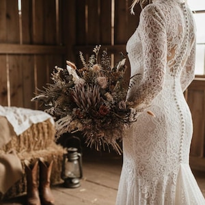Könnte beinhalten: Eine Braut in einem weißen Spitzenhochzeitskleid mit langen Ärmeln und hohem Ausschnitt, die einen Strauß Trockenblumen hält. Das Kleid hat einen Knopfverschluss hinten. Der Hintergrund ist eine rustikale Holzscheune.