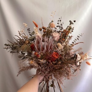 May include: A dried flower bouquet featuring a variety of textures and colors. The arrangement includes roses, protea, and other dried flowers in shades of orange, brown, and cream. The bouquet is held together with natural stems.