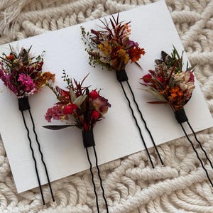 Puede incluir: Cuatro horquillas florales con horquillas negras. Cada horquilla presenta un pequeño ramo de flores secas en tonos rosa, rojo, naranja y amarillo, envuelto con cinta adhesiva negra. Las horquillas están dispuestas sobre una superficie blanca.