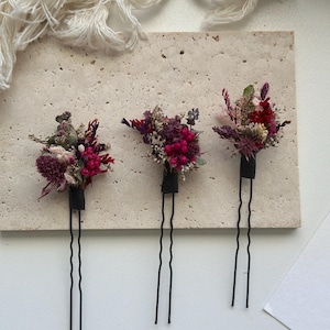Puede incluir: Tres horquillas florales con flores secas en tonos rojos, rosas y morados. Cada horquilla presenta un pequeño ramo sujeto a una base negra y un largo pasador negro ondulado. Las horquillas están dispuestas sobre una superficie clara y texturizada.