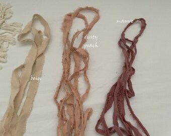 Puede incluir: Tres cintas de tela deshilachada de diferentes colores. Los colores son beige, melocotón polvoriento y malva.