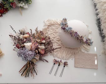 May include: A dried flower bouquet with a light purple hydrangea, pink roses, and other dried flowers. There are three hair pins with dried flowers on them, a dried flower crown, and a white round prop.