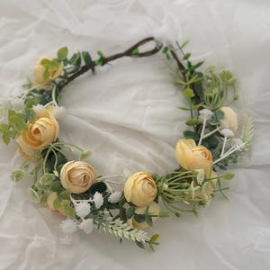 May include: A floral crown with pale yellow roses, green foliage, and white details. The flowers are arranged on a brown, twig-like base, creating a natural and delicate aesthetic. Suitable for weddings, festivals, or special occasions.