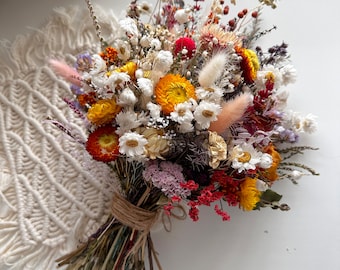 Pode incluir: Um buquê de flores secas com uma mistura de flores brancas, amarelas, laranjas e vermelhas. O buquê é amarrado com barbante e tem um estilo rústico e boêmio.