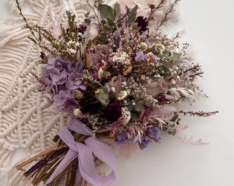 Könnte beinhalten: Ein Trockenblumenstrauß mit einer Vielzahl von Texturen und Farben. Das Arrangement umfasst lila Hortensien, burgunderfarbene Akzente und zarte weiße und rosa Blüten. Die Stiele sind mit Naturfaser und einem hellvioletten Band gebunden.