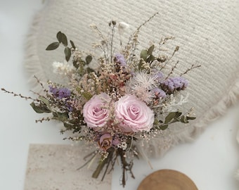 May include: A bouquet of dried flowers featuring two large, light pink roses as the focal point. The arrangement includes various dried flowers and greenery in shades of purple, white, and brown. The bouquet is tied together with natural twine.