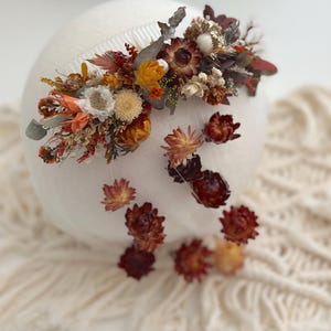 Puede incluir: Un peine para el cabello de flores secas con una mezcla de flores naranja, rojas y marrones. El peine está sujeto a un fondo blanco.