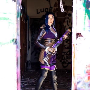Peut inclure: Une personne porte un costume de cosplay violet et marron avec un fusil dans une porte. Le costume comprend un haut violet, des accents marron et des leggings noirs. La porte est dans un bâtiment couvert de graffitis.