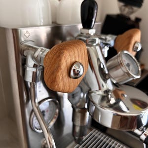 Op de afbeelding: Een roestvrijstalen espressomachine met opvallende lichtgekleurde houten knoppen met een textuur in de houtnerf. De machine bevat een ronde drukmeter, een metalen lekbak en een zwarte hendel, alles uitgevoerd in een metalen afwerking.