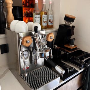 Könnte beinhalten: Eine Lelit Espressomaschine aus Edelstahl mit Holzknöpfen, einem Manometer und einem Siebträger. Vier weiße Keramiktassen stehen oben auf. Daneben befindet sich eine schwarze Kaffeemühle mit einem Holzdeckel und einer kleinen schwarzen Tasse.