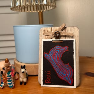 May include: A rectangular piece of paper featuring a black background with a stylized map of Peru outlined in red and blue lines. The word Peru is written in red text. The paper is held by a metal clip on a wooden board with a jute bow.