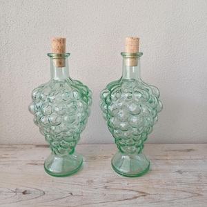 May include: Two clear light green glass bottles shaped like clusters of grapes. Each bottle features a textured surface with rounded embossed spheres and a narrow neck topped with a cylindrical cork stopper, standing on a circular glass base.