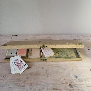 May include: A rectangular, distressed cream-coloured wooden box with a hinged lid containing two decks of playing cards and a "How to Play" instruction booklet. A single playing card showing the Queen of Clubs is placed in front of the box.