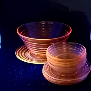 May include: A set of vintage glass bowls and saucers. The bowls and saucers have a ribbed design and a warm orange hue. The set includes a large bowl, a smaller bowl, and matching saucers. The glass appears to glow against a dark background.