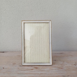 May include: A rectangular picture frame with a white and gold beaded border. The frame is empty, with a light-coloured backing visible. The frame is displayed on a light-coloured wooden surface.