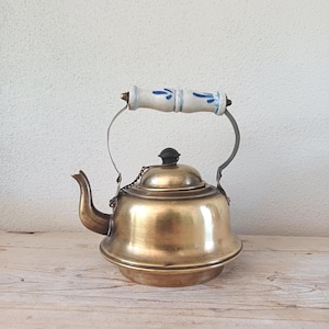 May include: A vintage brass kettle with a ceramic handle decorated with blue floral patterns. The kettle has a curved spout, a black lid knob, and a metal handle. It rests on a wooden surface, suitable for a kitchen or dining area.