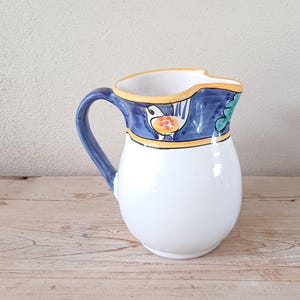 May include: A white ceramic jug with a blue and yellow floral design. The jug has a handle and a spout.