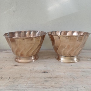 May include: A pair of matching copper bowls featuring a scalloped rim and a distinct vertical wave pattern along the exterior. Each bowl is supported by a circular pedestal base, showcasing a warm metallic finish with subtle variations in tone.