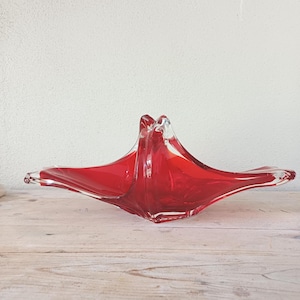 May include: A striking red glass bowl with a distinctive starburst design. The bowl features a wide, open form with pointed edges, crafted from transparent red glass. It is presented on a light-coloured wooden surface.