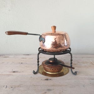May include: A copper fondue pot with a wooden handle and lid, on a black metal stand. A smaller copper burner with a spoon sits on a brass plate below. The pot has a hammered texture, typical of vintage cookware.