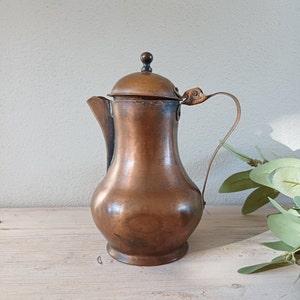 May include: A copper teapot with a lid and a handle. The teapot has a rounded body and a slightly flared spout.