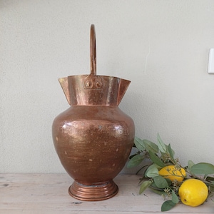 May include: A copper pitcher with a hammered finish and a curved handle. The pitcher has a wide mouth and a rounded body. It is sitting on a wooden surface.